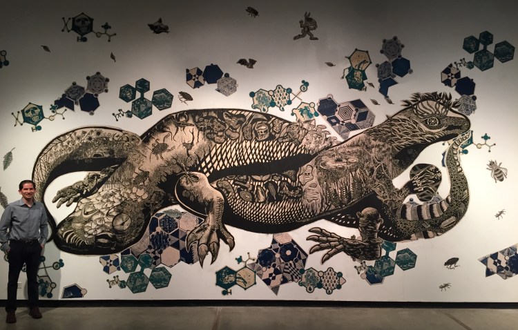 Dean Saúl Jiménez-Sadoval stands by a printmaking mural made by artists Mazatl and Kill Joy at Arte Americas