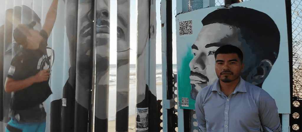 Issac stands in front his images on the mural with QR codes which send people to his story on YouTube. Credit Julio Cesar Ortiz-Franco.
