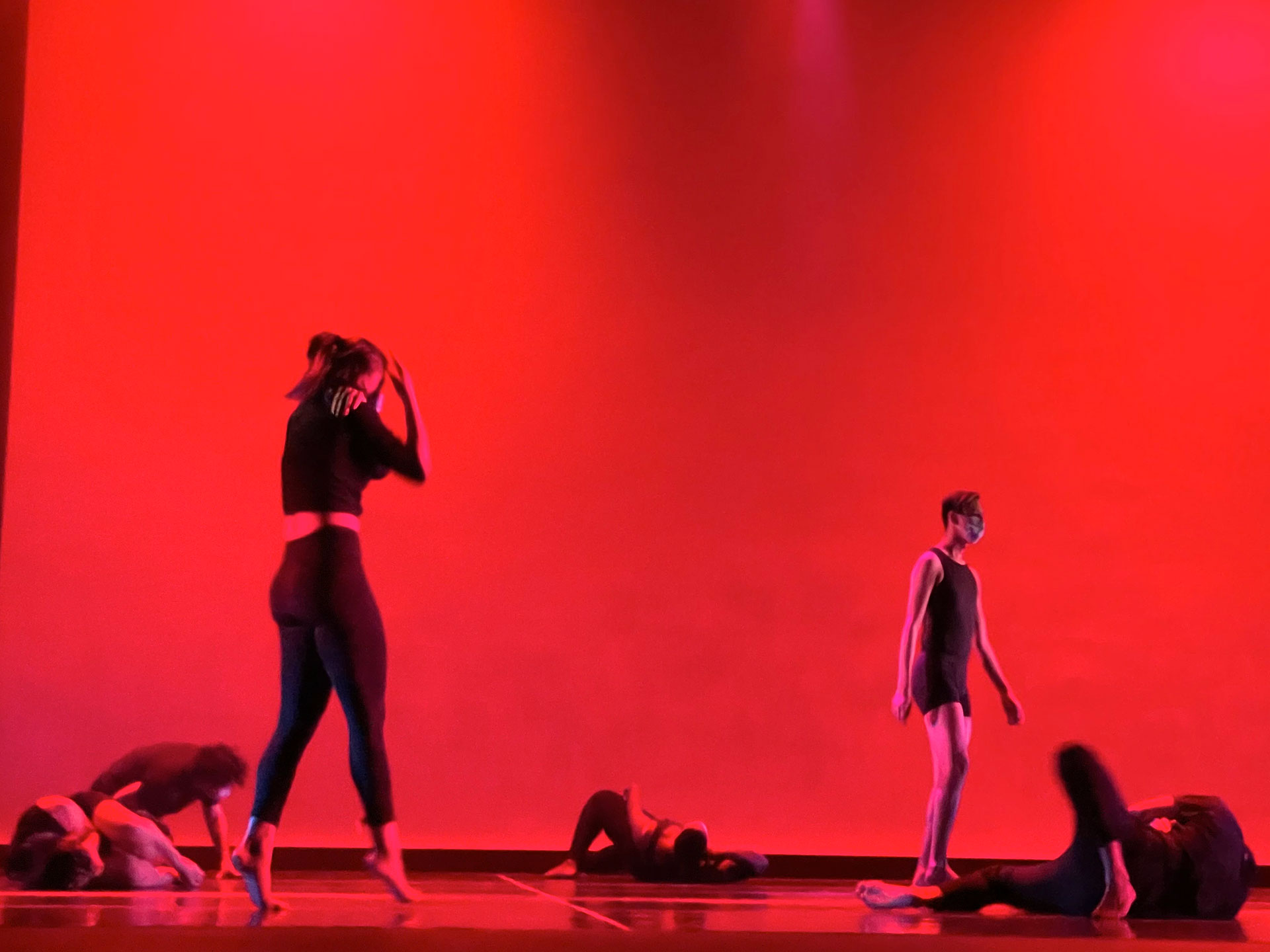 Two dancers roam the stage while other dancers lay down on stage in front of a brilliant red background.