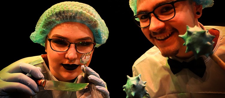 A woman and a man, loosely dressed as dentists, peer into your mouth. The woman is holding a knife and fork while the man holds sticks with horned balls on the end.