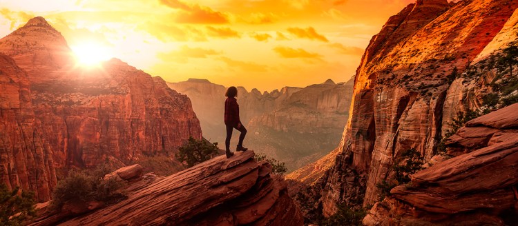 A woman stands on a red rock overlooking a canyon as the sun sets.