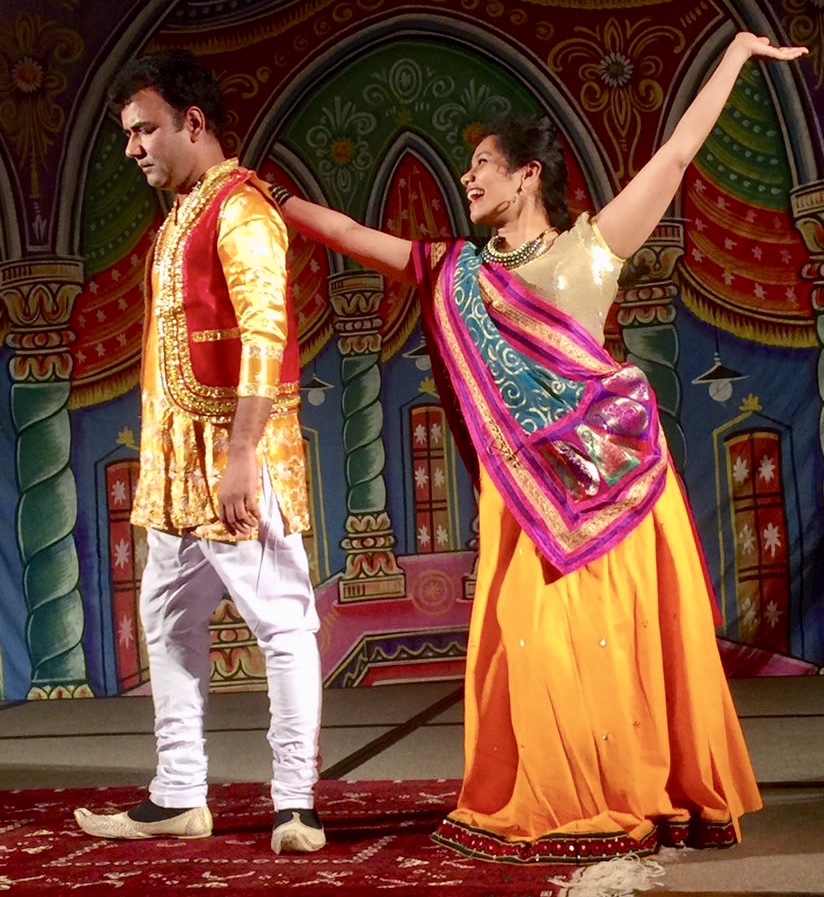 Dr. Devendra Sharma in a gold and red traditional outfit performs in a nautanki with a yellow dress and a purple, teal and gold wrap.