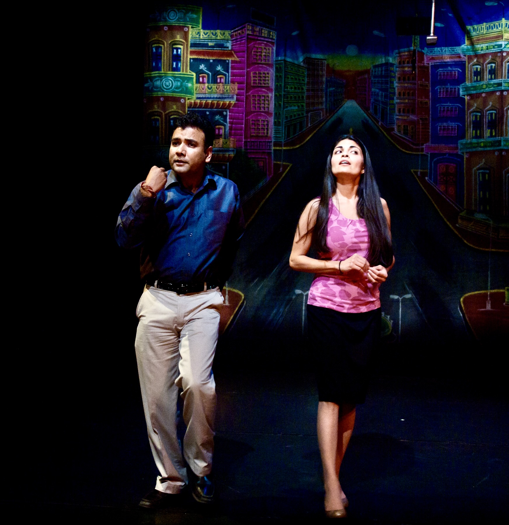 Dr. Devendra Sharma and a fellow actress "walk" in front of a colorful street backdrop in contemporary clothing.