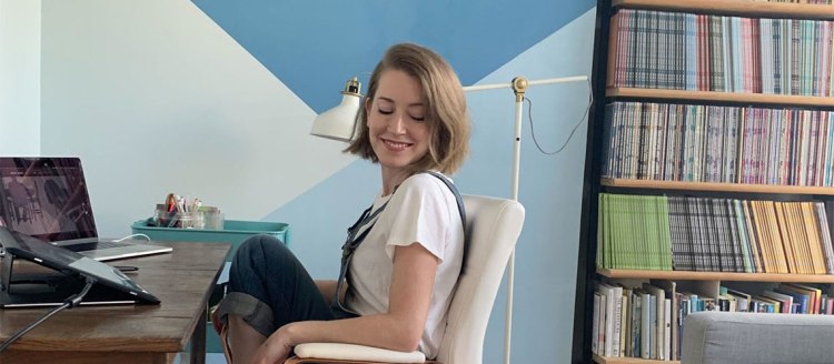 Kristen Radtke, author of the graphic memoirs “Seek You: A Journey Through American Loneliness” (2021) and “Imagine Wanting Only This” (2017) sits in a chair in her office in front of a computer, but she is looking down. A bookshelf is behind her.