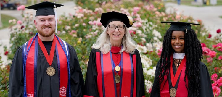 Undergraduate Dean's Medalist Steven Hensley, Dr. Honora Chapman, and Graduate Dean's Medalist Audia Dixon are wearing their commencement regalia and wearing their Dean's Medals in front of the rose garden with blooms of white, pink, yellow and red.