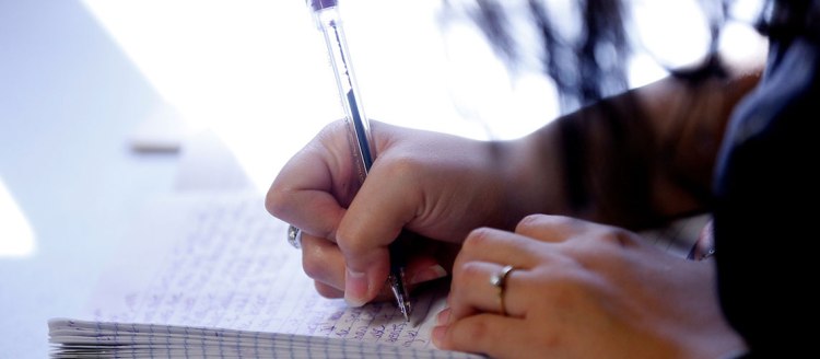 A woman's hand with rings on her fingers grasps a pen tightly as she writes in a notebook.