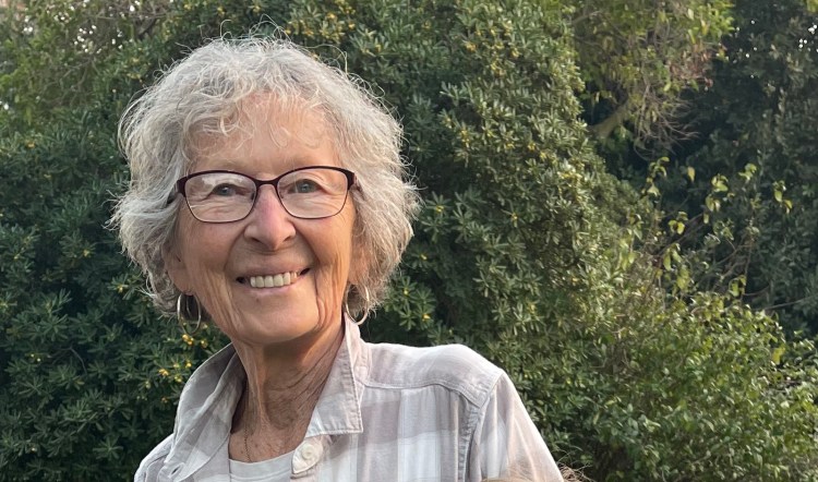 Frances Levine smiles in a loose light gray colored shirt in front of trees.