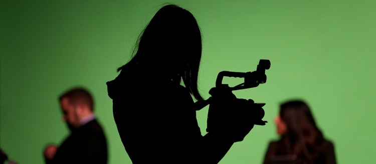 Silhouetted photographer holds a camera in front of a green screen with two journalist ready to be filmed.