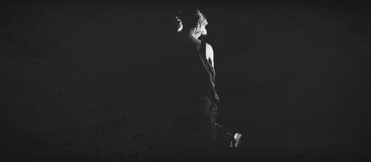 A woman with dramatic backlighting on black background, looks down in this black and white photo.