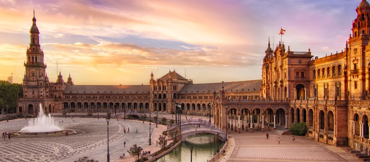 The Plaza de España in Seville is an large stone building, promenade and fountain with a foot bridge over a canal during sunset.