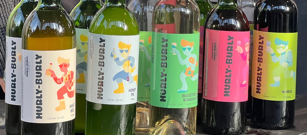 Hurley-Burly wine lables on five types of wine bottles. The logo is a vector image of a guy with sweeping hair holding a bottle and walking.