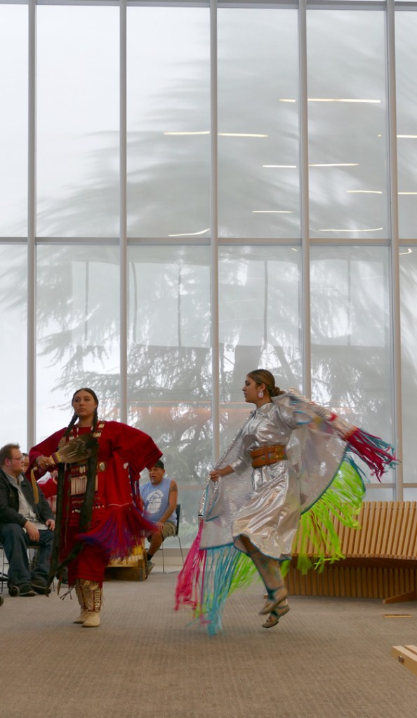 Indigenous dancers perform, one wearing red and the other whitish silver traditional outfits dance in front of a large opaque window.