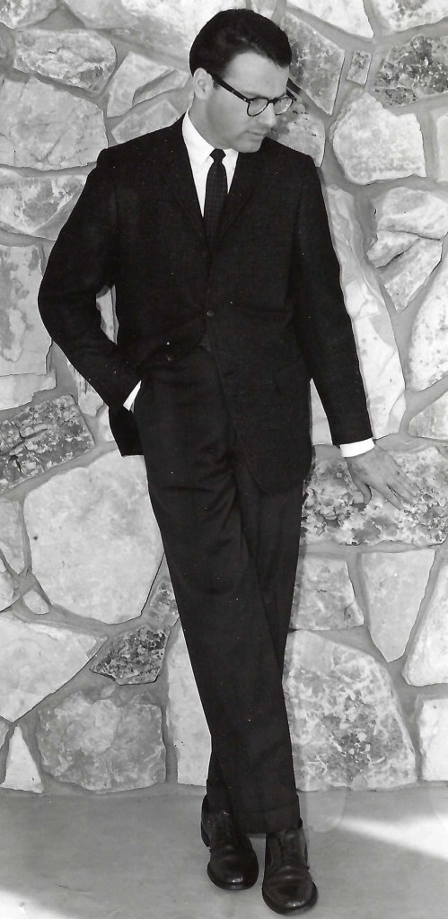 A black and white photo Ralph Shabazian in a black suite in front of a tall stone wall.