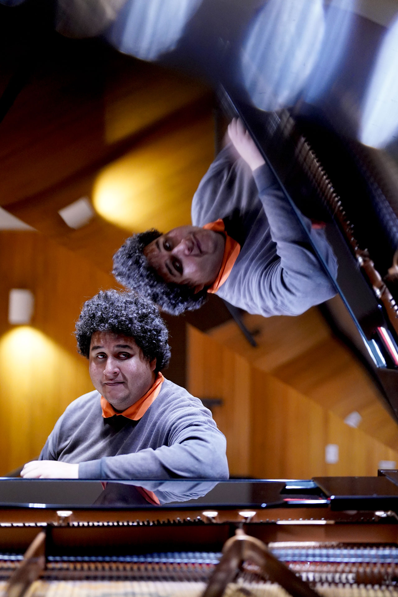 Maurissio poses at the grand piano with the lid open. His reflection is seen in the bottom of the lid.