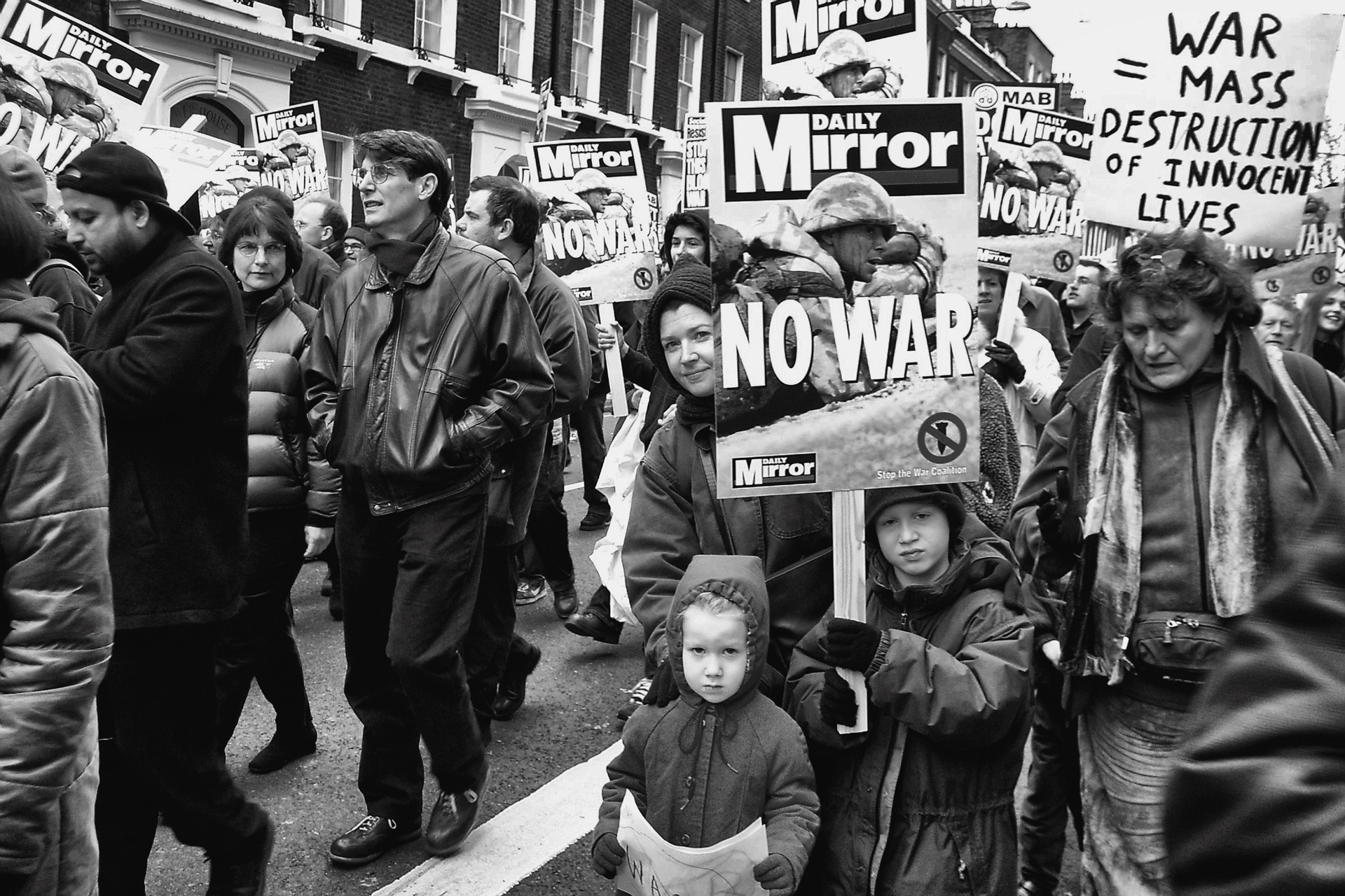 The image is black and white monochrome of a group of protestors walking down a city street. Brick buildings are in the background and we see protestors, including young children, holding printed signs that read "Daily Mirror - NO WAR" and a photo of a soldier with blood running down his face. One protestor has a handmade sign that reads "War=Mass destruction of innocent lives."