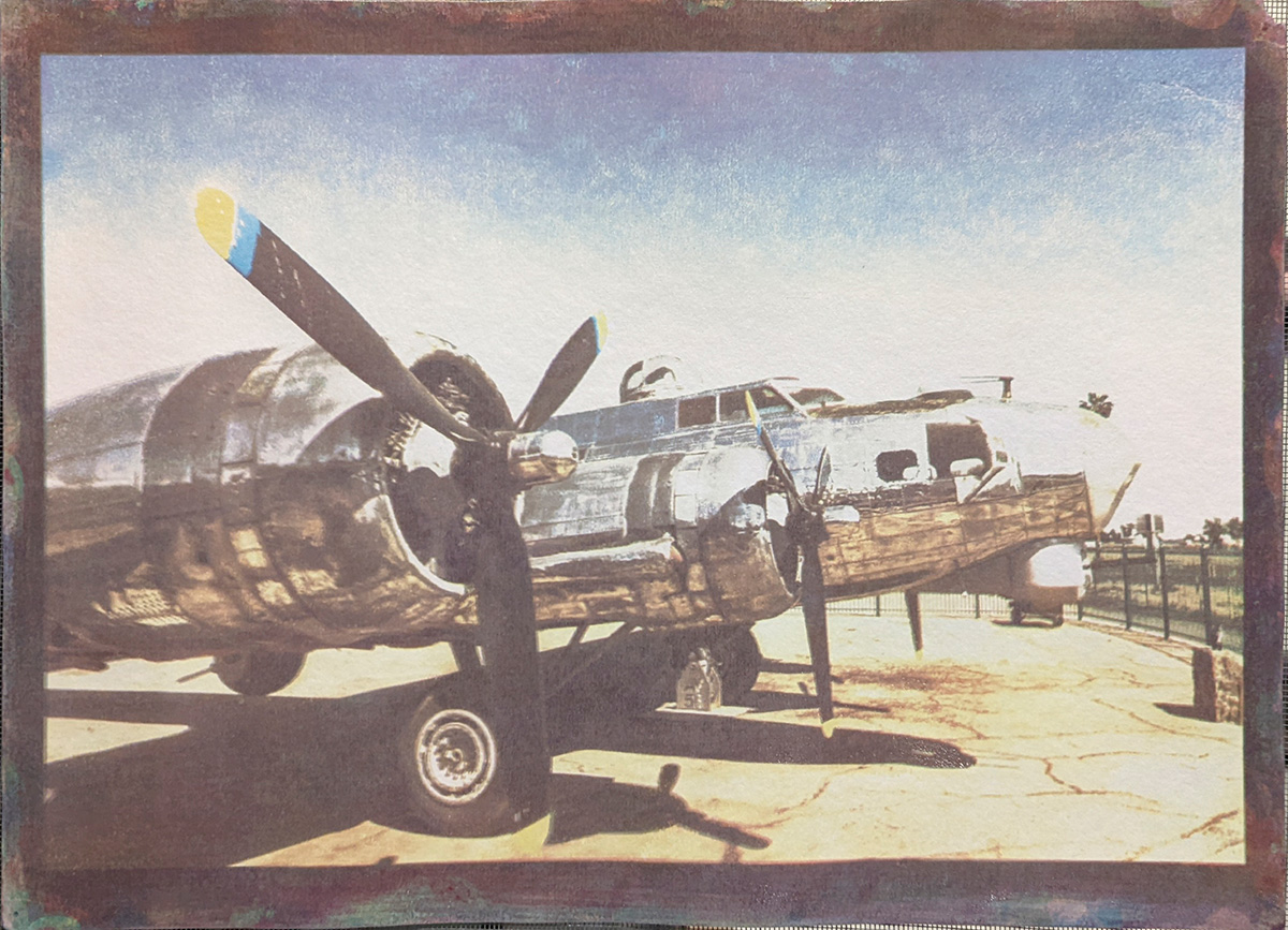 A colored side profile of a chrome B-17 bomber parked on a lot surrounded by a black metal fence. The propellers of the plan are painted yellow and blue at the tip though the plane looks worn. The photograph is framed by a black border from the paper which the image is printed on.
