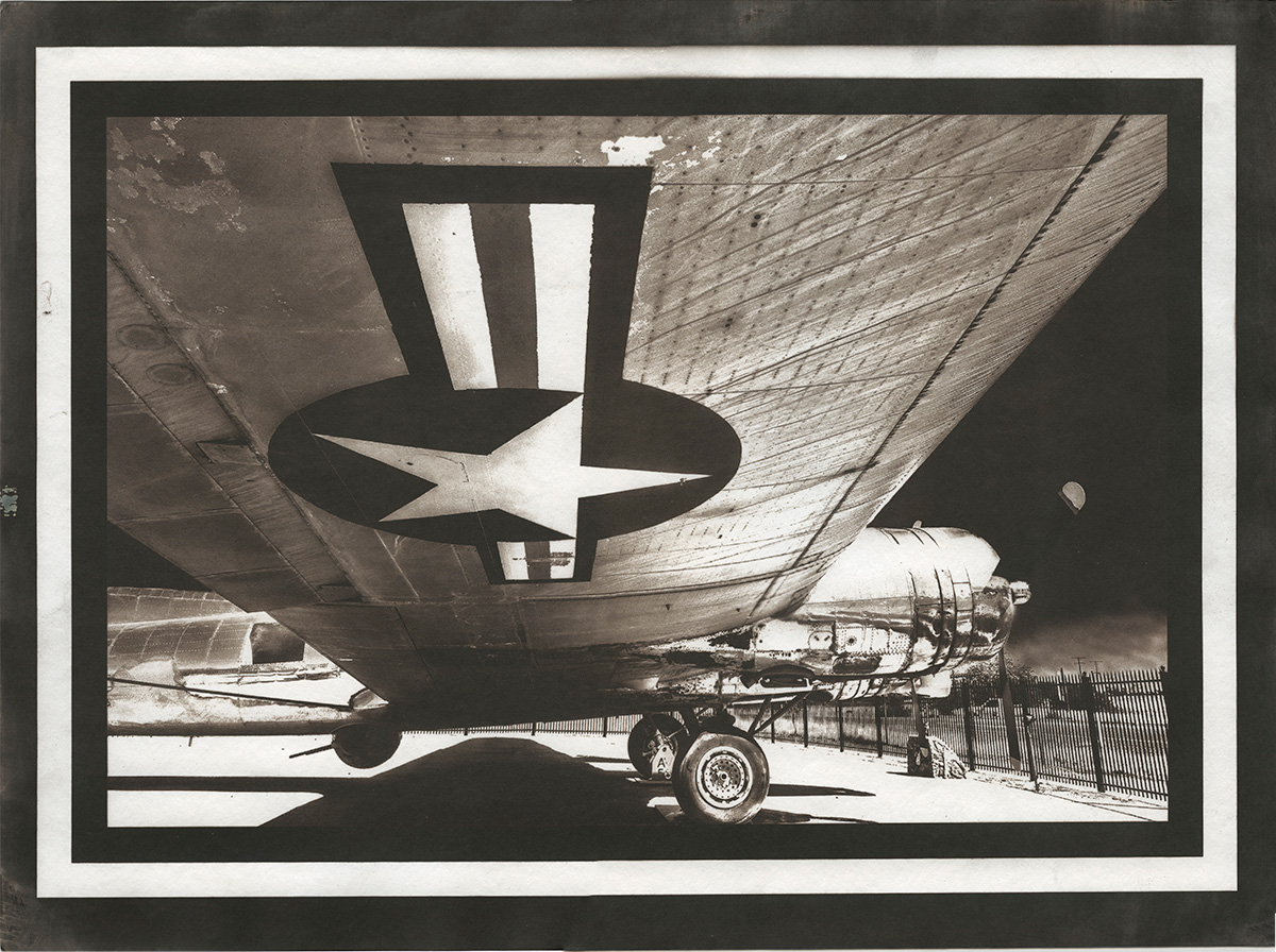 The image is a monochrome black and white with a B-17 bomber framed from underneath its right wing with the insignia of the American Airforce. The sky that is visible is dark, though there is strong sunlight on the shining, metallic bomber.