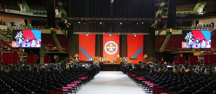 A wide shot from the back of the College of Arts and Humanities commencement shows students sitting listening as a speak talks on stage in the distance. Large screens flank the stage which has a red and blue backdrop with the campus insignia.