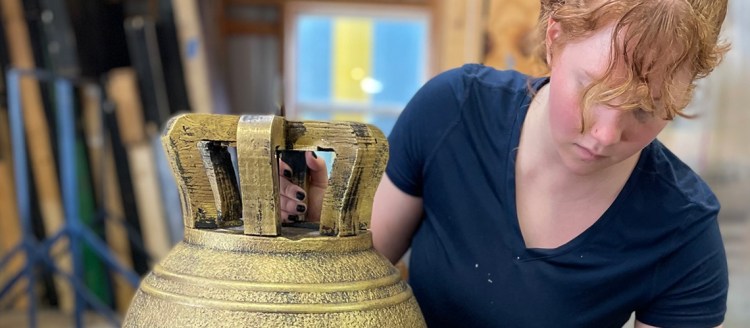 Cecilia Fisher adding gold paint to a prop bell. She is wearing a. blue shirt.