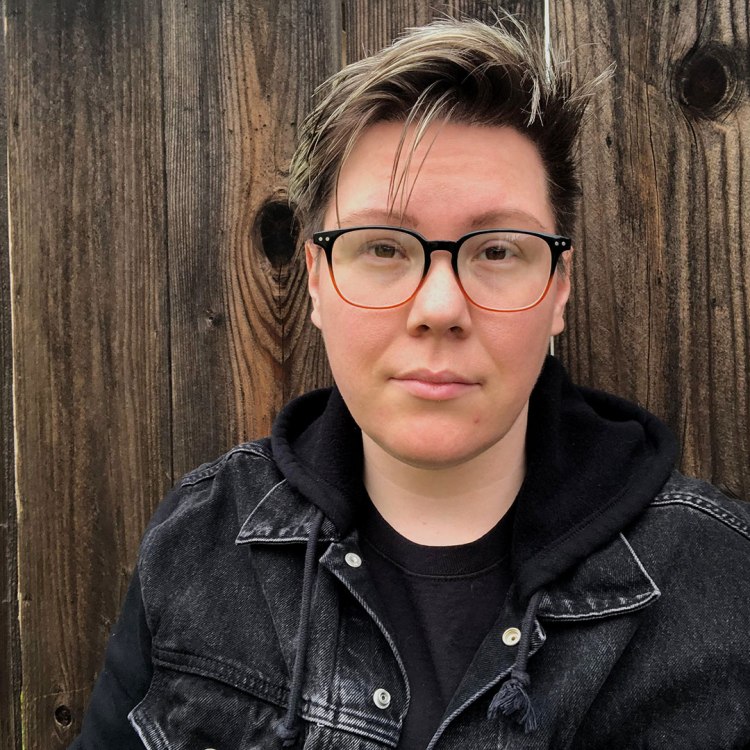 Recent Fresno State graduate Icarus Bailey who cross references Fresno State's herbarium specimens with words used by the Chukchansi tribe. He stands in front of a wood fence and is wearing a black denim jacket with a hood, and a black shirt.