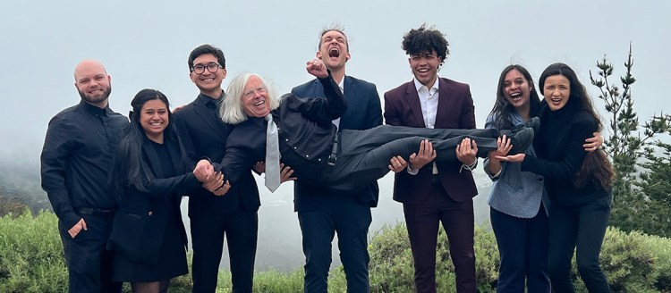 The Barking Bulldogs hold up their coach Dr. Douglas Fraleigh. Everyone is dressed in business casual dress and they stand on top of a mountain, behind them we can see trees and a foggy valley.