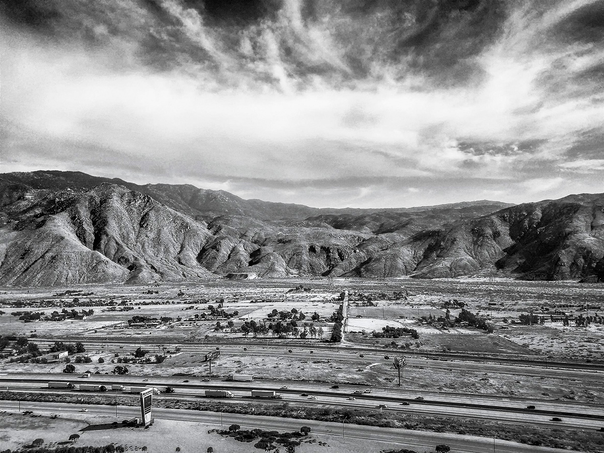 A black and white image of a valley. Closer to the viewer we see a highway, and a little further on some houses. Beyond the houses is flat land with small vegetation, which suddenly turns into moderately sized mountains.