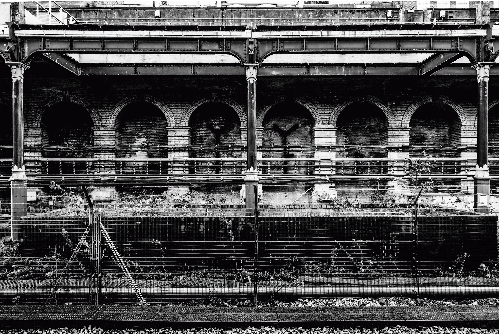 A crisp black and white image of a train platform. We see a part of the tracks at the bottom of the image. There is a fence separating the tracks from an overgrown platform. A metal structure holds up a roof above the platform, and further back is a large brick building supported by arched columns.