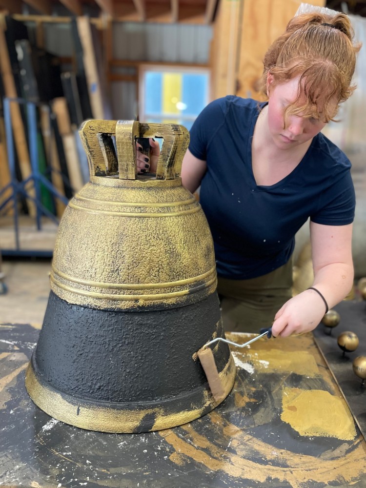 Cecilia Fisher adding gold paint to a prop bell. She is wearing a blue shirt and wood panels can be seen stored in the background. She holds the bell steady with her right hand and holds a paint roller in her left.