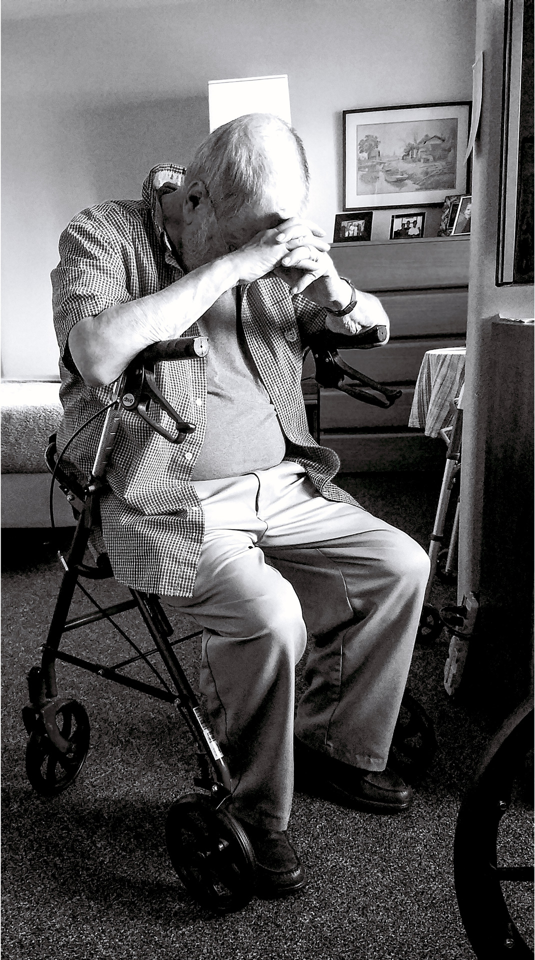 A black and white image of an old man with thinning white hair. He has a beard and glasses, as well as a watch on his left wrist. l He wears a t-shirt under a short sleeve, button up flannel, and closed toed slippers. He is slightly to the left of the photo sitting on a walker with wheels. He has his hands together interlocking fingers, and his head faces down and rests on his fingers. In the background there is an obscured table and bed, as well as a dresser with photographs on it and a painting above it. A light is right behind the old man's head.
