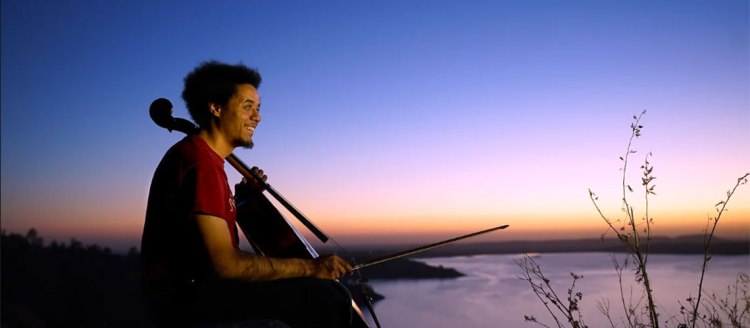 Dalton Morris plays his cello outside overlook a lake at sunset.