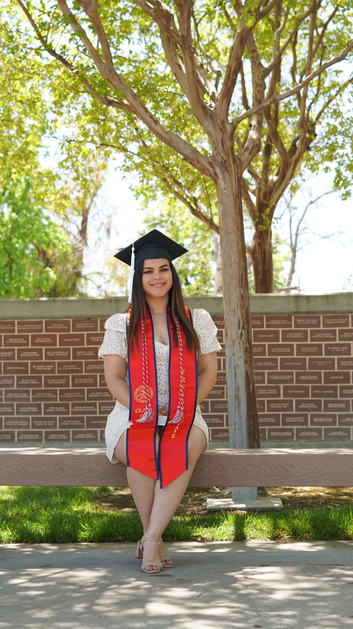 Nayeli Guerrero sits on a bench with her Fresno State stole and grad cap in front of a brick wall with trees.