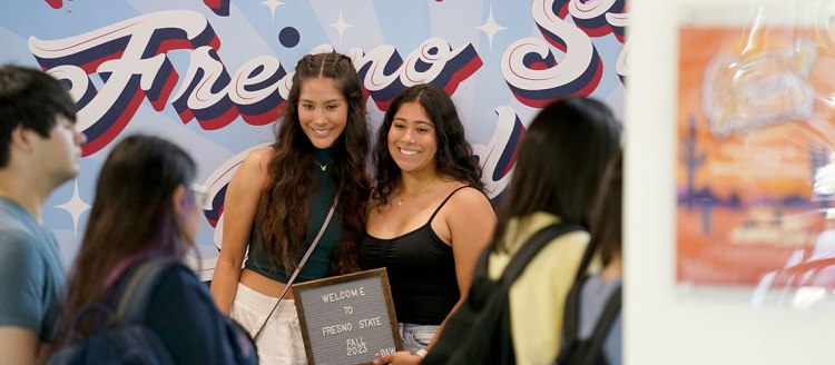 Students stand in front of a light blue backdrop with "Fresno State" in large white, blue and red script lettering. Two young ladies are holding a small sign that says "Welcome to Fresno State Fall 2023"