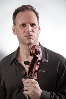 A man stands with his face and upper body framed. He is wearing a grey shirt, and is holding the neck of a cello in front of him