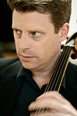 A close up photo of a man, with his face and upper chest being framed in the photo. He has brown hair and is looking to the left of the viewer. He has a cello and his hand moving on the fingerboard.