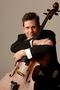 A man in a black tuxedo is sitting with his cello. He is wrapping both of his arms around the neck of the cello, is leaning on the cello, and is smiling at the camera.