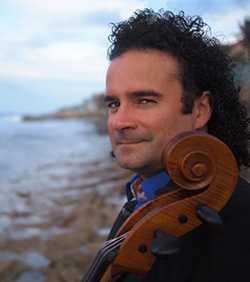 A man stands on the coastline with his cello. The headstock of the cello is visible, and we see that the man is on a beach. He has curly black hair.