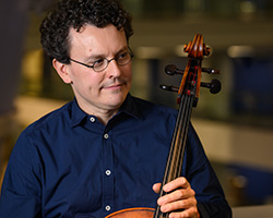 A man in a navy blue button up shirt holds his cello. He has black curly hair, round black glasses, and is looking off to the right.