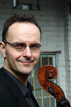 A man stands in front of a grey brick building. He has black hair, black glasses, a grey button up shirt, and a black jacket. He is looking at the camera and holding his cello in front of him.