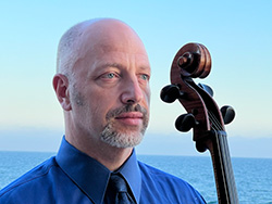 A man stands with his cello, the top of the neck and the headstock are visible, and positioned next to his face. The man wears a blue collared shirt with a tie, and the ocean and blue sky can be seen in the background.