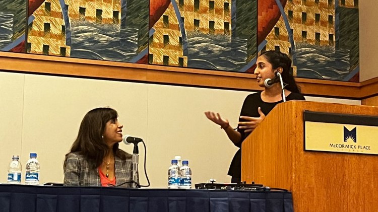 Dr. Veena Howard sits at a panel and looks up at Fresno State Student Shailey Shah who is at a podium and presenting on Jain food practices.