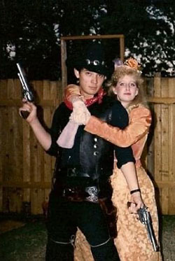 Actor Daniel Swartz in an all-black cowboy outfit with a 6-shooter pistol in each hand while actress Laura Robinson hugs him from behind in a peach western era dress.