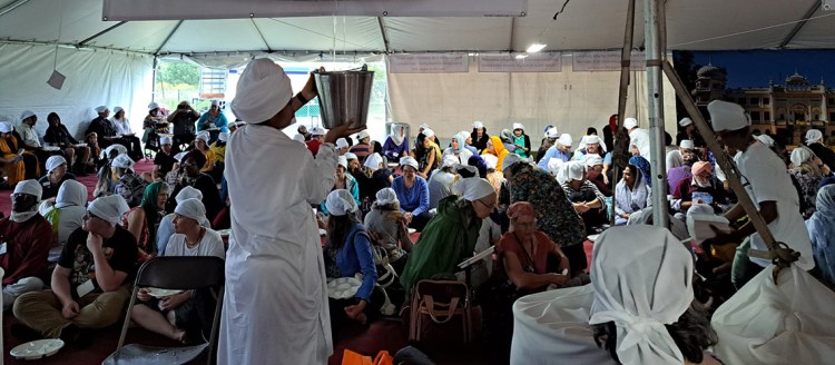 A white tent is filled with people sitting on the ground. A Sikh man in dressed in all white and a head covering holds up a bucket full or curry. He uses a ladle to give curry to any person who wants it. Everyone is wearing white head scarves and have taken their shoes off, as is religious custom.