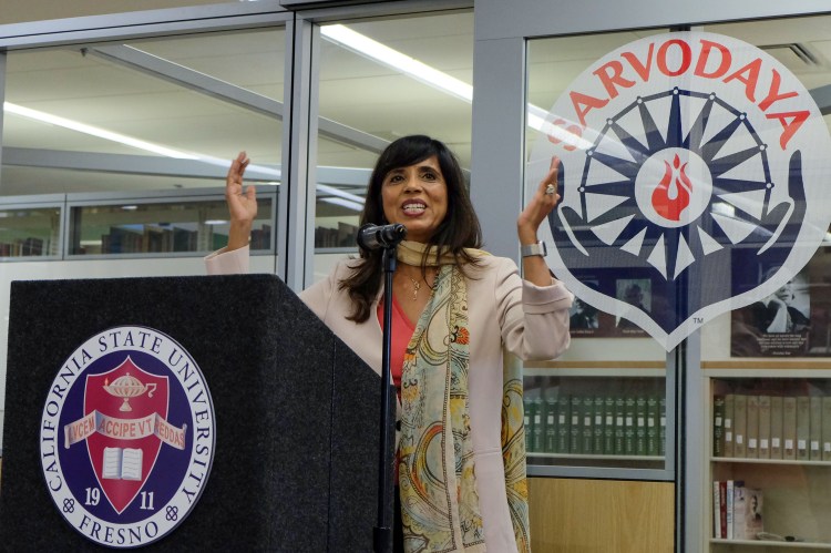 Dr. Veena Howard speaks at a podium branding the Fresno State logo. She has her hands raised and behind her is the M.K. Gandhi Center. Its logo can be seen which is two cupped hands holding a wheel with a flam in the center.