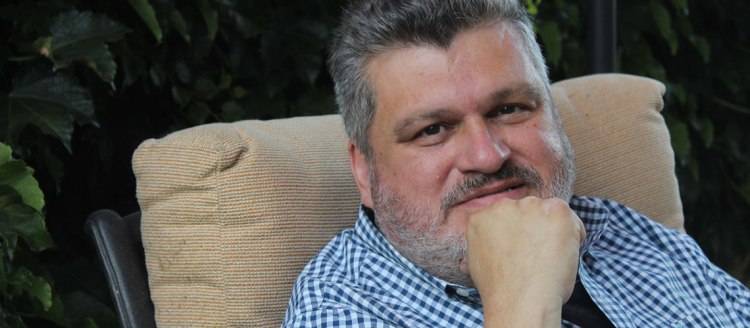 Ruben Navarrette in a blue and white checkered shirt sitting outside with his fist to his chin.