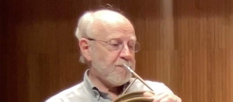 A man in a white collared shirt stands while playing a valveless French horn. He has white hair, glasses, and a beard.