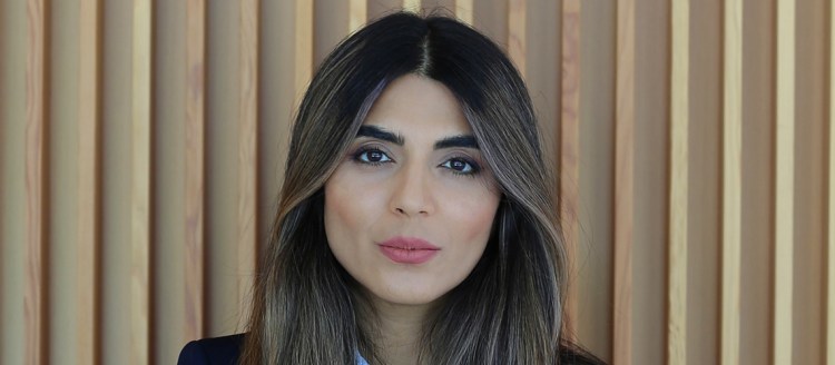 New faculty member Nasrin Golshany stands in front of a wall overlayed with thin wood running parallel across. She has black hair that is dyed lighter, and is wearing a navy blue suit with a white dress shirt.