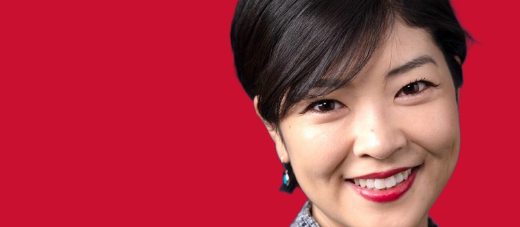 A woman's face is in frame. She has short cut hair, a turquoise earring, red lipstick, and is smiling with her head slightly tilted to the left. The background is a Fresno State red.