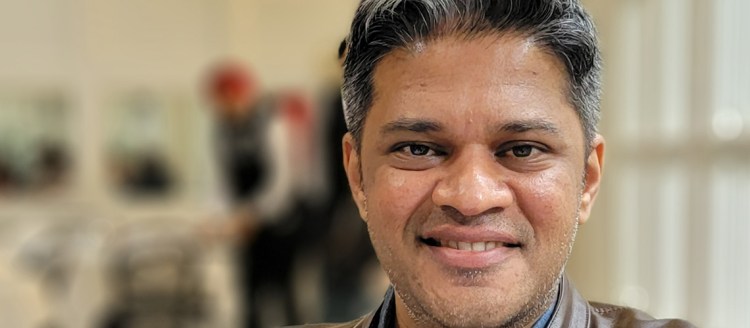 A man stands with his face in frame and focused, and the background blurred. He is smiling and has greying black hair. There are people in the background behind him. He is wearing a denim collared shirt and a brown leather jacket.
