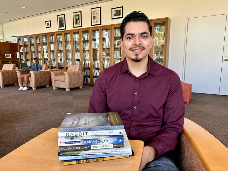 Luis Granados Torres sits in a chair with a stack of books on a table in front of him. A large bookshelf and chairs fill the background.