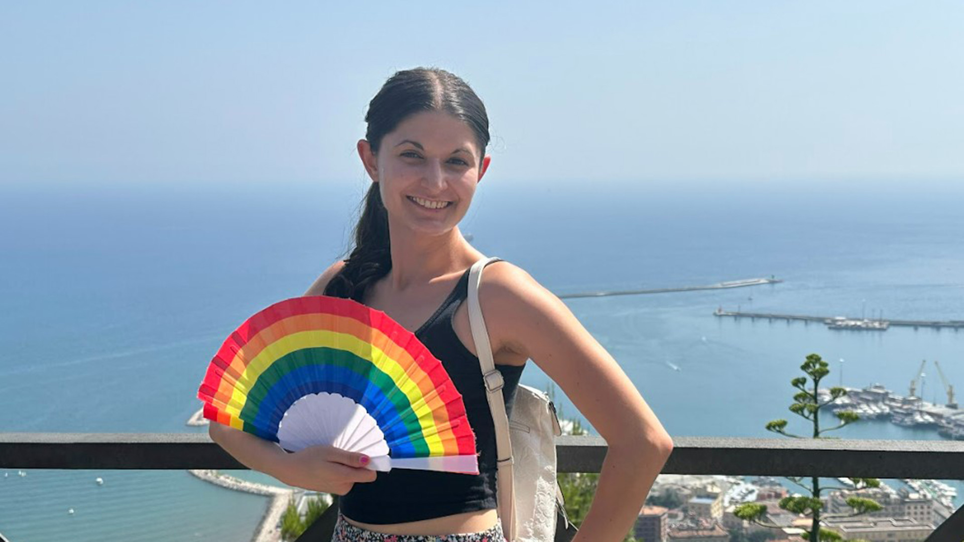 A woman standing with a rainbow fan in her hands, standing on a ledge over looking the edge of a coastal city.